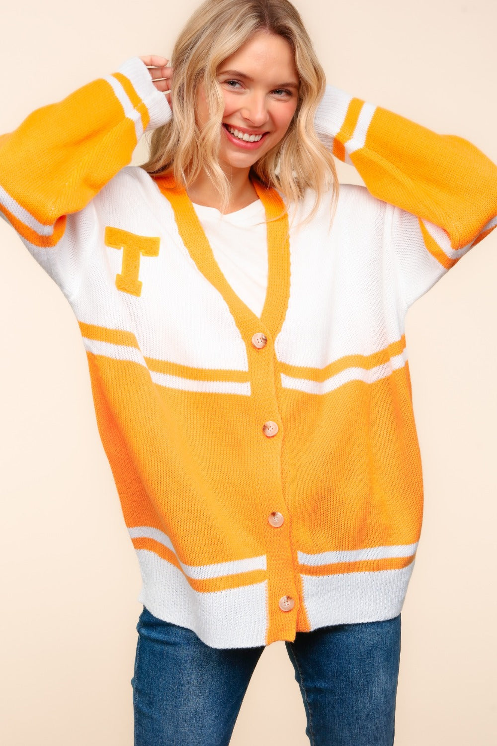 A smiling person with long blonde hair wears the Haptics V Neck Button Down Letter Patch Cardigan in bright orange and white. It features button details and a T patch on the chest, with their hands playfully over their head, paired with casual jeans.