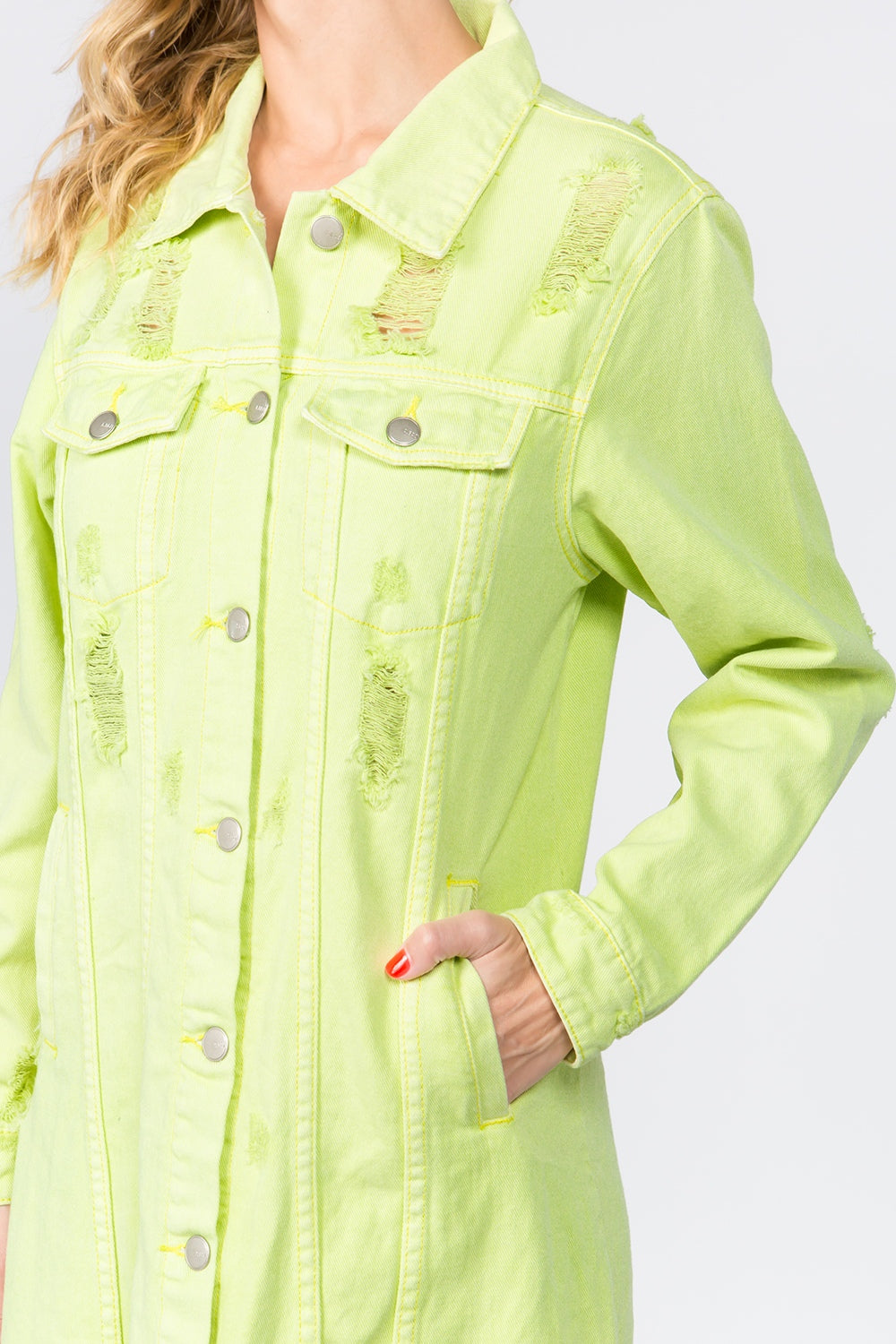 Someone models the American Bazi Button Up Distressed Denim Dress in light green, featuring silver buttons and chest pockets. With one hand in their pocket and their head out of view, the mystery is intact, complemented by their red-painted nails.