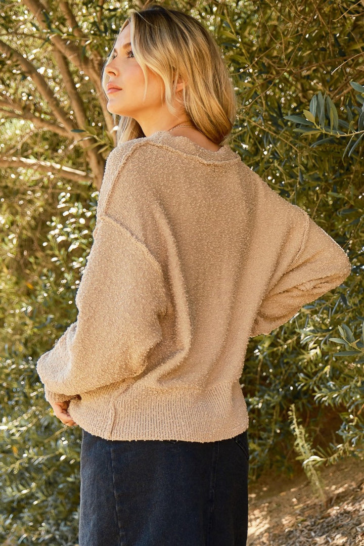 A person with long blonde hair stands outdoors, slightly turned away. They are wearing the distinctive And The Why Button Down Popcorn Sweater Cardigan paired with black pants. Greenery and sunlight create a natural, serene backdrop.