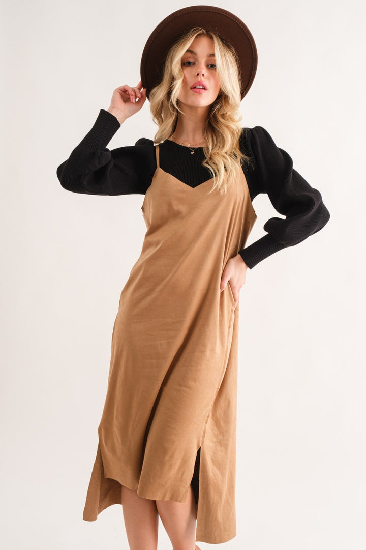 A person models the And The Why Faux Suede Cut Edge Slit Cami Midi Dress in tan over a black long-sleeve top, accessorizing with a wide-brimmed brown hat. The dresss knee-length hem and sophisticated style create a casually elegant look against a plain white background.