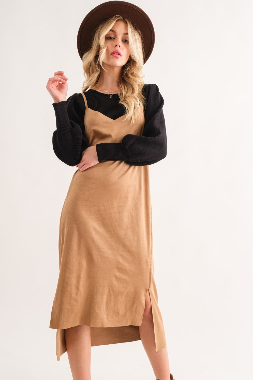 A woman with long, wavy blonde hair models the And The Why Faux Suede Cut Edge Slit Cami Midi Dress in black, complemented by a wide-brimmed brown hat. She exudes sophistication while posing against a white background, gently holding her dress to reveal its side slits.