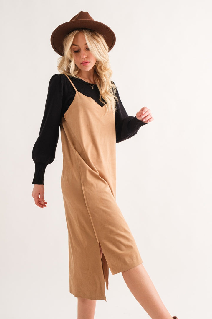 A woman with long blonde hair exudes sophisticated style in the And The Why Faux Suede Cut Edge Slit Cami Midi Dress, paired with a black long-sleeve top and a brown hat. She stands against a plain white background.