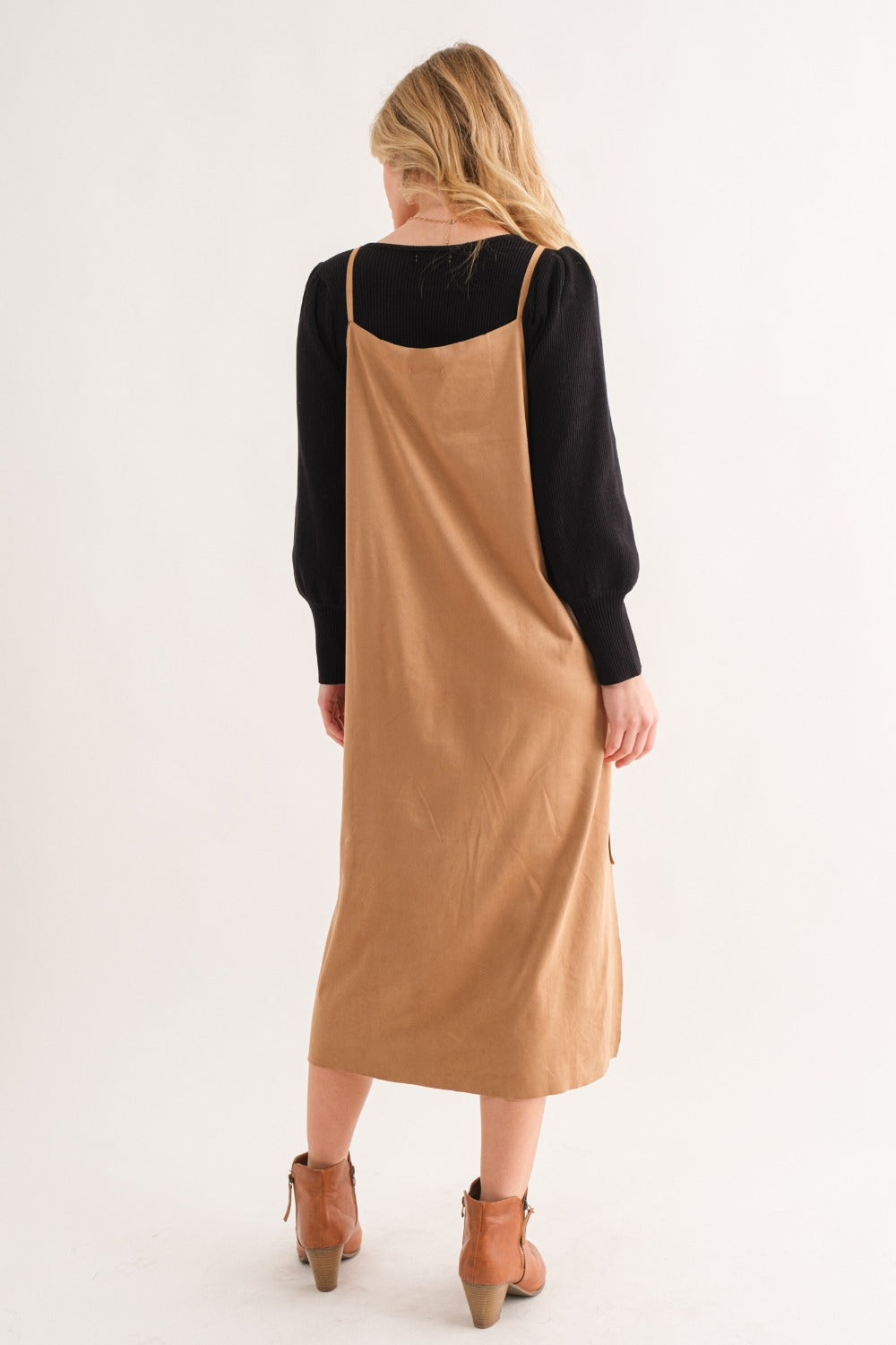 The image depicts a person with long blonde hair, facing away, in an And The Why Faux Suede Cut Edge Slit Cami Midi Dress in brown, layered over a long-sleeve black top. They are standing on light brown heels set against a neutral background.