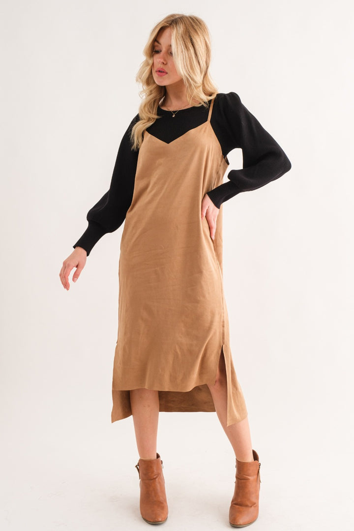 A person with long blonde hair gracefully poses against a plain white background, wearing the elegant And The Why Faux Suede Cut Edge Slit Cami Midi Dress in light brown, layered over a black long-sleeve shirt and paired with brown ankle boots.