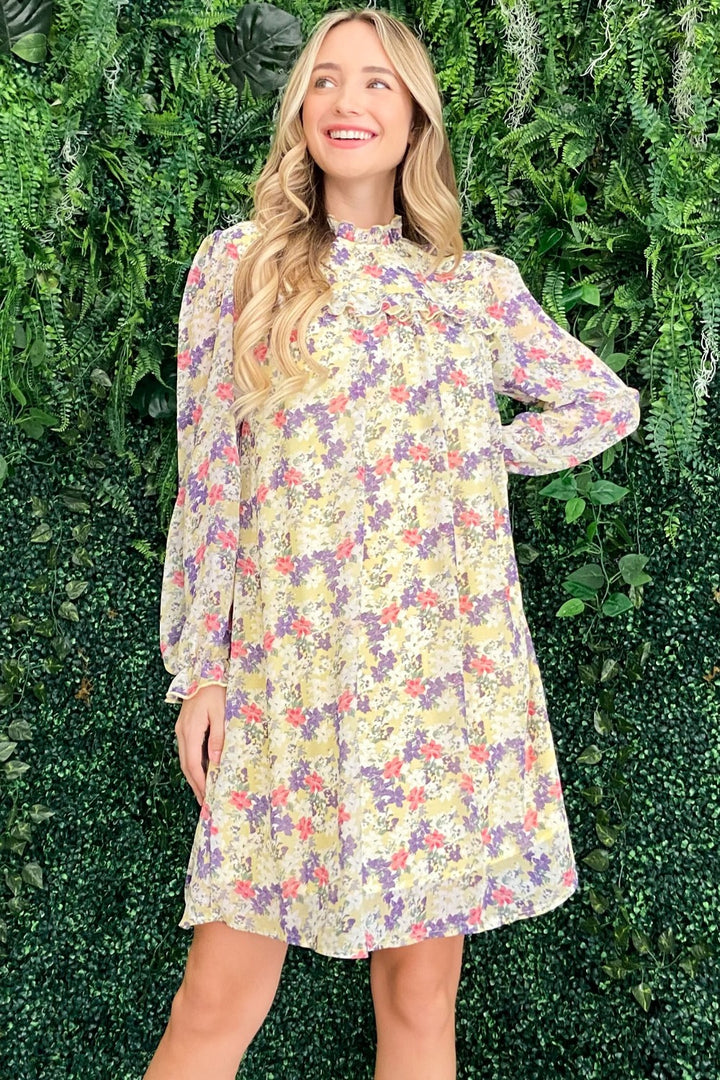 A woman in an And The Why Floral Mock Neck Flounce Sleeve Dress smiles, her long blonde hair flowing, as she stands in front of a lush green wall filled with plants, posing with one hand on her hip.