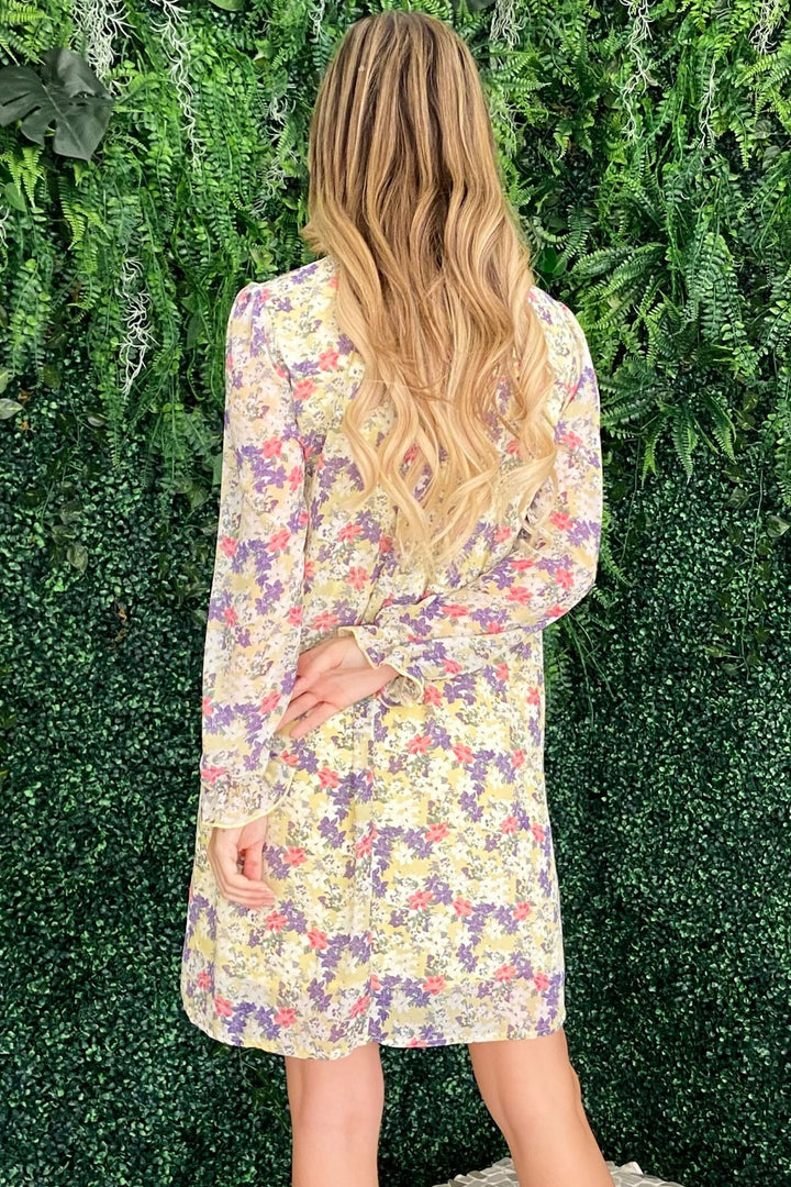 Facing a wall of lush green foliage, a person with long, wavy blonde hair wears the And The Why Floral Mock Neck Flounce Sleeve Dress. Their left hand is on the back of their head, and their right hand rests on their lower back.