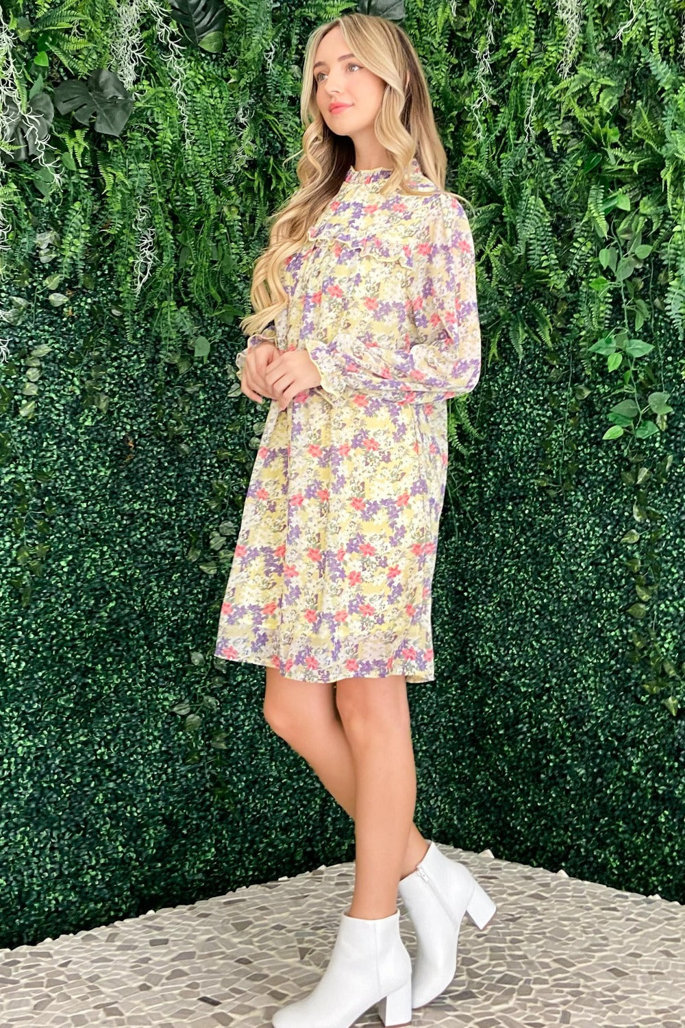 A woman in the And The Why Floral Mock Neck Flounce Sleeve Dress and white ankle boots stands on a mosaic tile floor, her long hair cascading over her shoulders, set against a backdrop of lush green foliage.