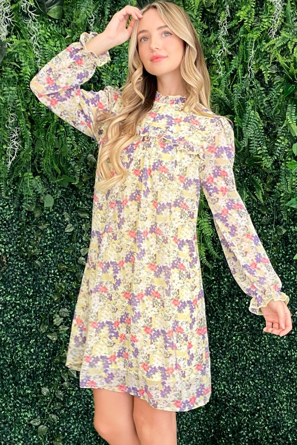 A woman stands before a leafy backdrop, modeling the And The Why Floral Mock Neck Flounce Sleeve Dress. Her long blonde hair flows in loose waves as she gently smiles, one hand near her face.