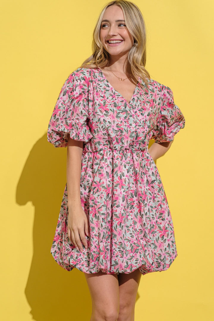 Someone in an And The Why Full Size Floral Surplice Puff Sleeve Dress, featuring a feminine pink floral pattern and cinched waist, stands against a bright yellow background, smiling as their long, wavy hair cascades over their shoulders.