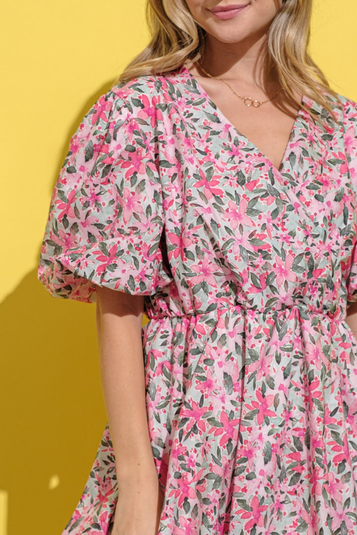 A woman in an And The Why Full Size Floral Surplice Puff Sleeve Dress poses against a bright yellow background, featuring pink flowers and green leaves, puffed sleeves, and a gathered waist. Her hair is loose, she wears a delicate necklace, and her face is partially visible.