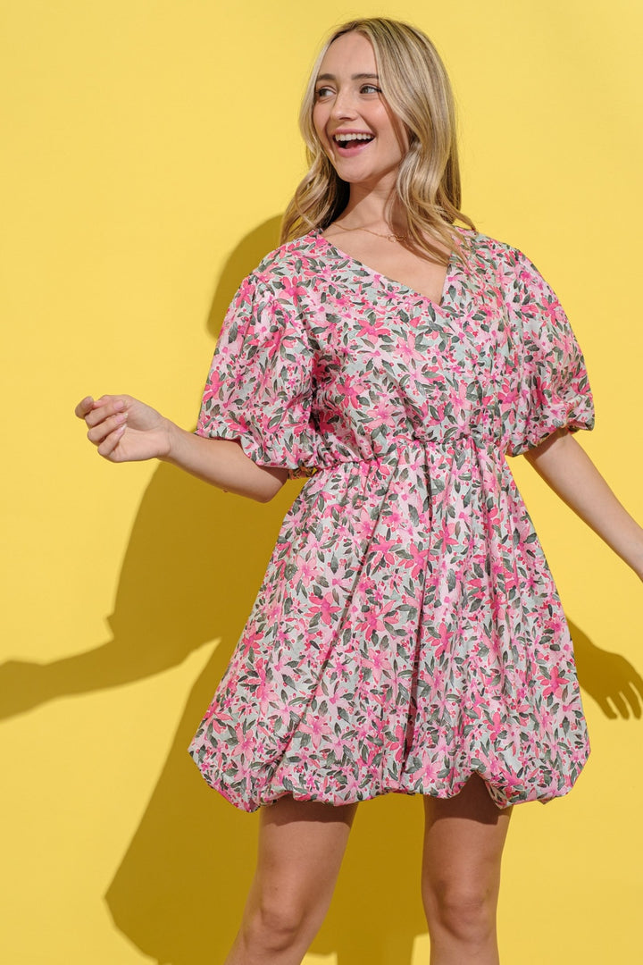 A person in an And The Why Full Size Floral Surplice Puff Sleeve Dress, adorned with pink and green floral patterns, smiles against a bright yellow background. Their hair cascades down as they gently extend their arms.