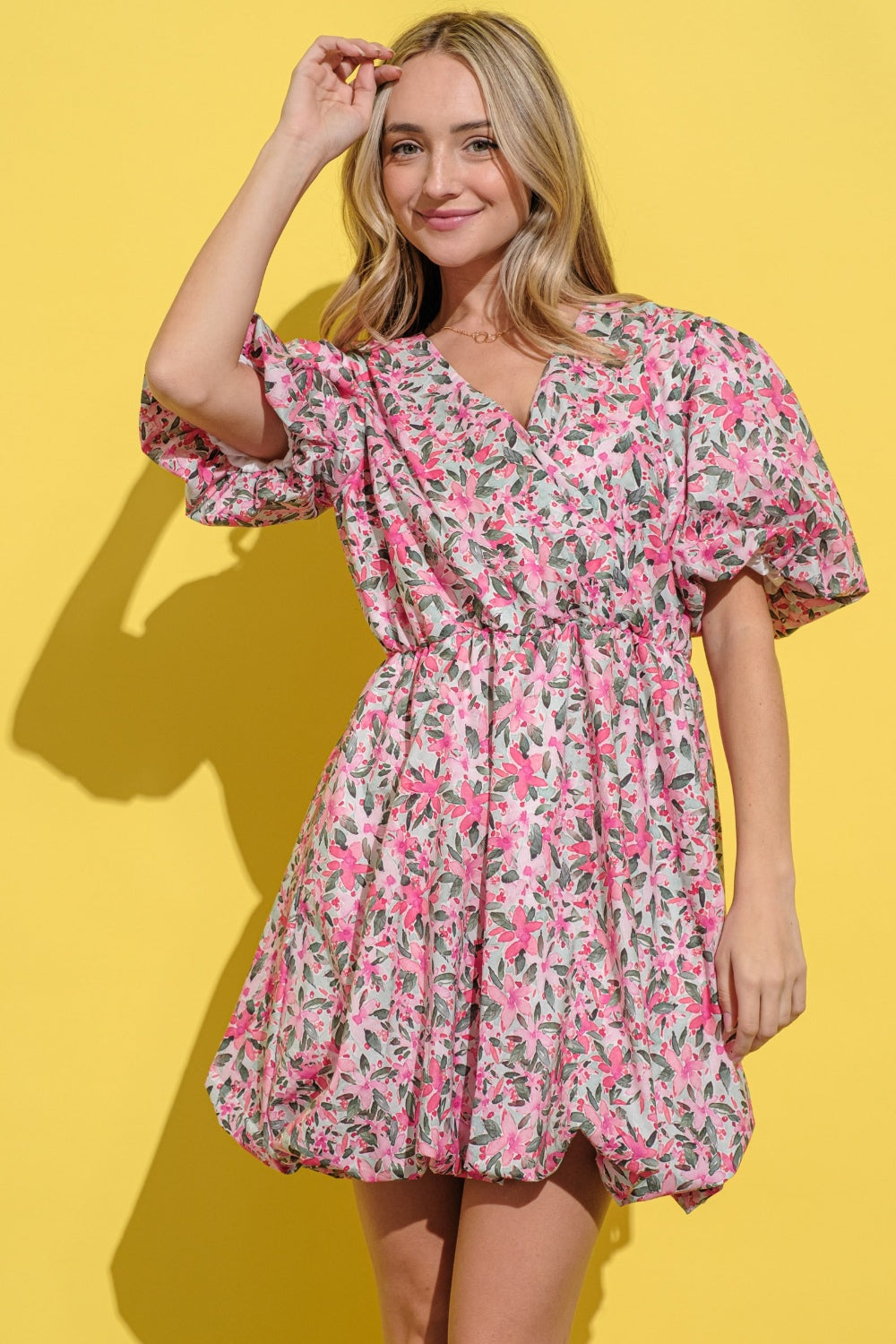 A woman wears the And The Why Full Size Floral Surplice Puff Sleeve Dress, featuring a bubble hem and pink floral print, as she stands against a yellow background, smiling with one hand in her hair.