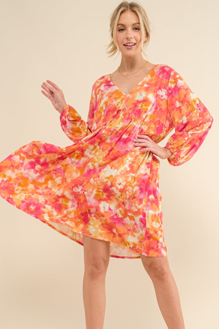 A woman is wearing the And The Why Full Size Printed Tie Back Long Sleeve Dress, featuring a fashionable and elegant floral pattern in vibrant shades of pink, orange, and white. This dress gracefully flows above the knee as she smiles and poses against a neutral background.