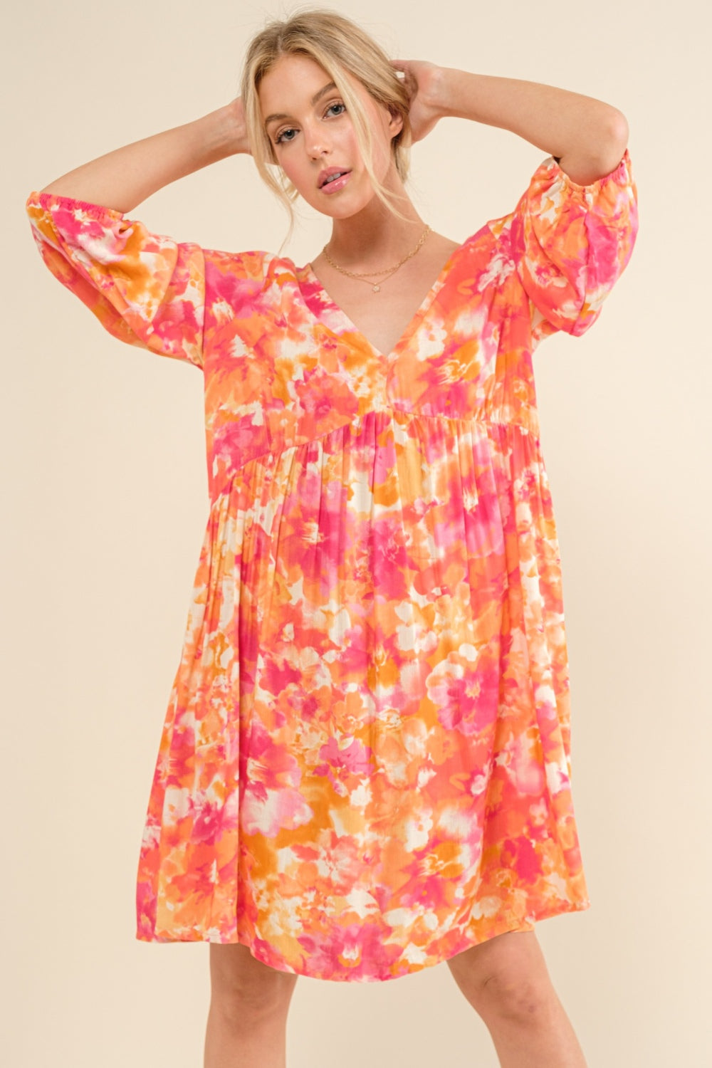 A person poses in the And The Why Full Size Printed Tie Back Long Sleeve Dress, showcasing its flowy, fashionable design adorned with vibrant floral patterns in shades of pink, orange, and yellow. This elegant piece features a V-neckline and puffed sleeves, all set against a plain beige background.