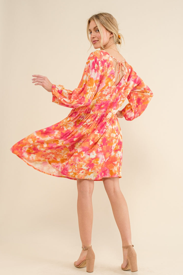 A woman twirls in the And The Why Full Size Printed Tie Back Long Sleeve Dress, which is fashionable and elegant with a flowing skirt. The dress showcases vibrant hues of pink, orange, and white. She pairs it with beige heels, and her loosely styled hair enhances the playful yet sophisticated vibe.