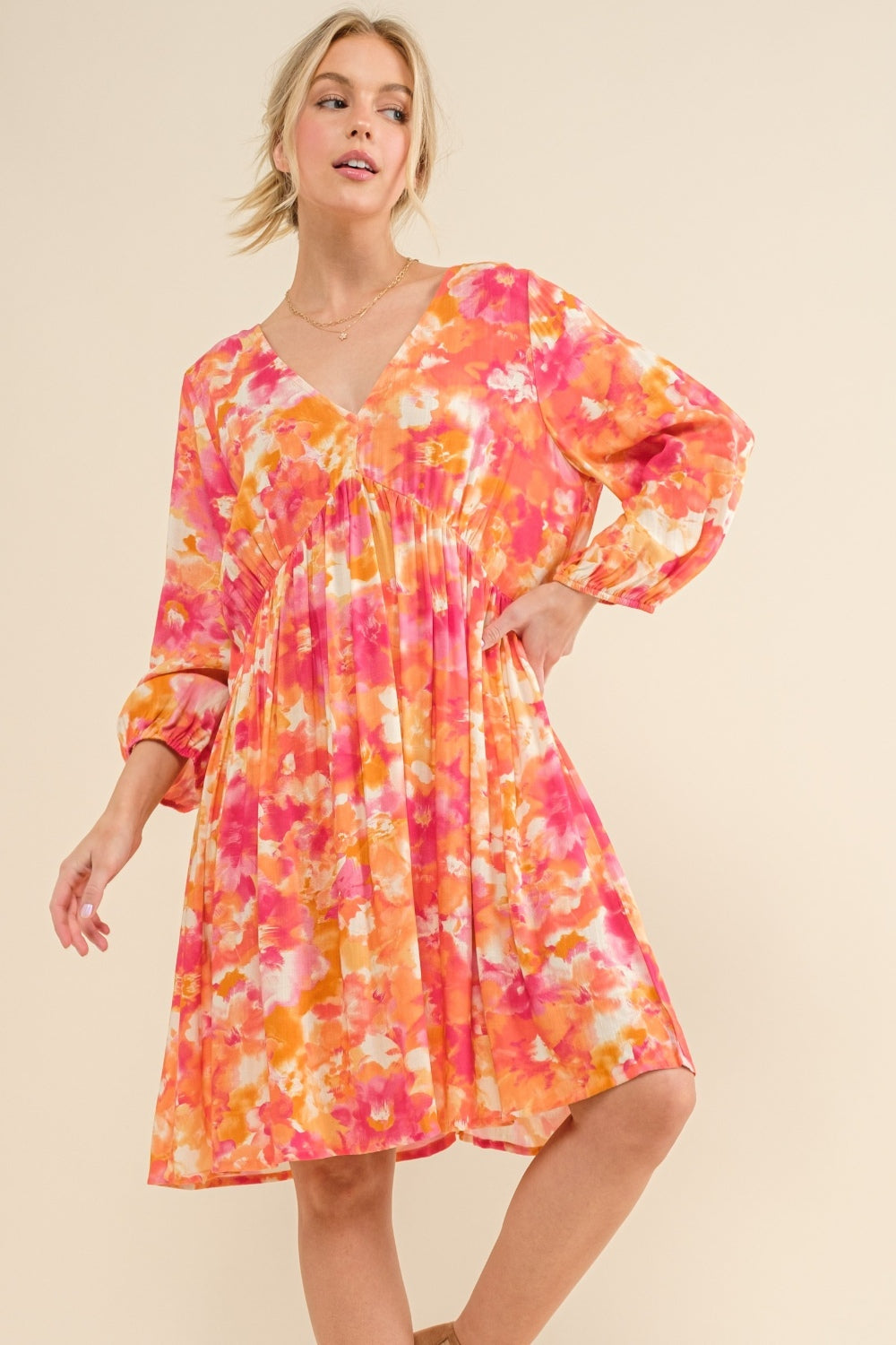 A person wearing the And The Why Full Size Printed Tie Back Long Sleeve Dress, showcasing a fashionable floral pattern in shades of pink, orange, and yellow. The dress features an elegant V-neck and flared sleeves, making it stand out beautifully against the plain, light background.