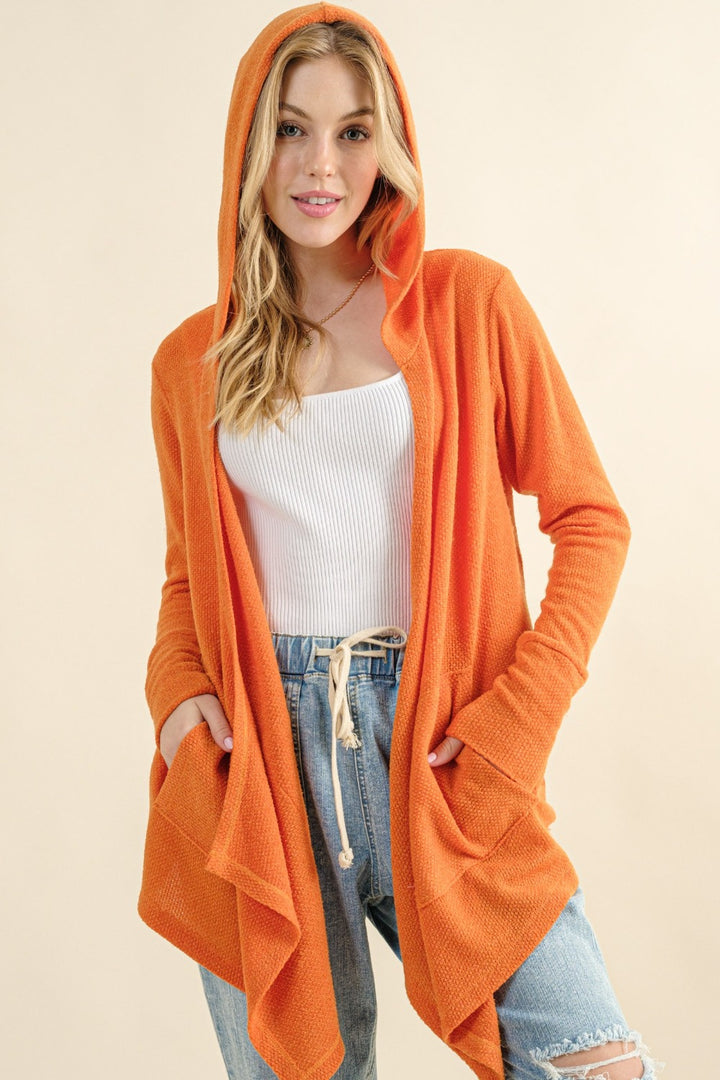 A person with long hair is wearing the cozy And The Why Full Size Thermal Hooded Open Front Cardigan in orange over a white tank top, paired with blue jeans. They have their hands in the cardigan's pockets, standing against a neutral background.