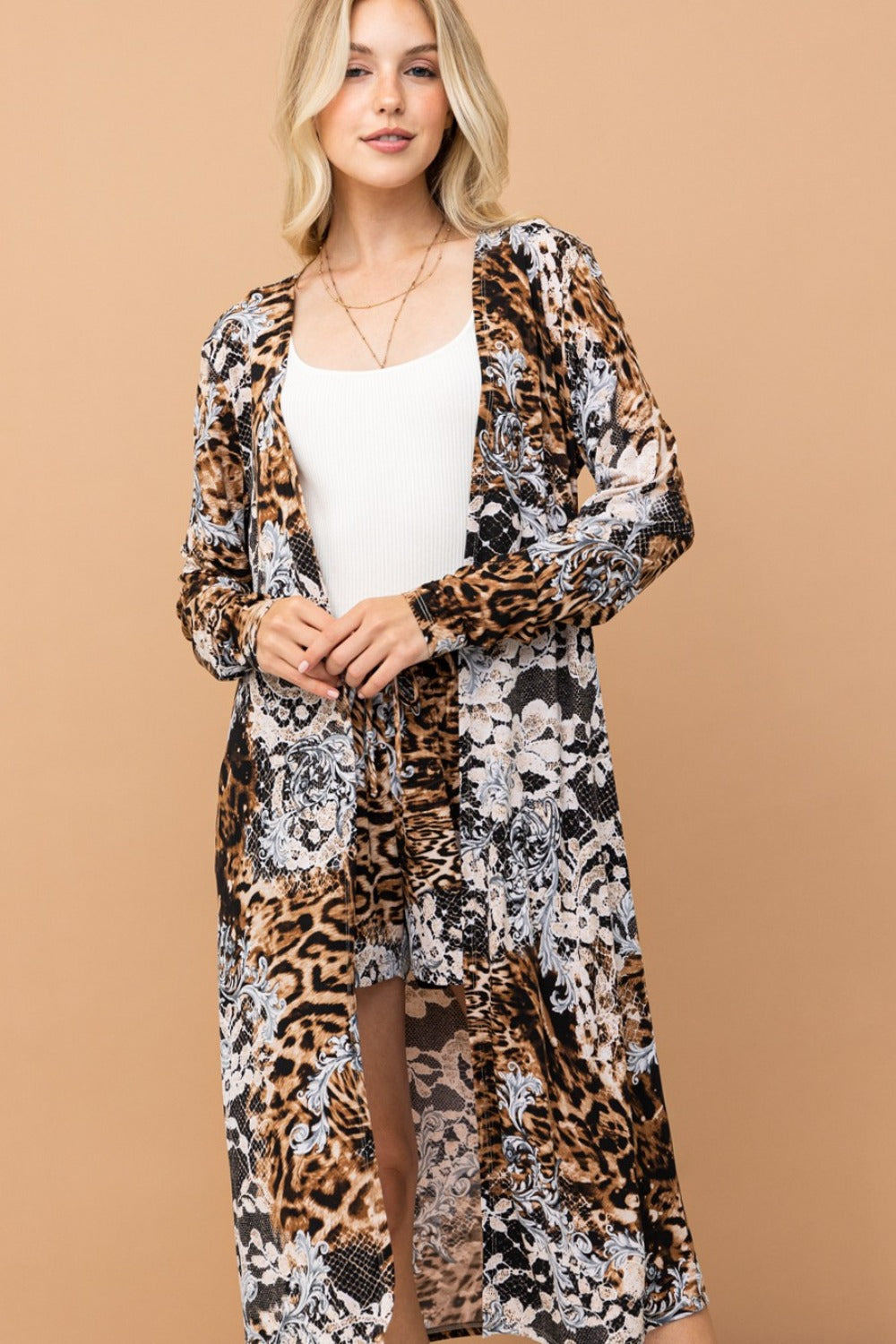 A woman is pictured standing against a plain beige backdrop, wearing the And The Why Leopard Kimono Open Front Longline Cardigan, which features a mix of animal and floral prints. Beneath it, she has on a white top paired with shorts. Her long blonde hair cascades down as she smiles softly.