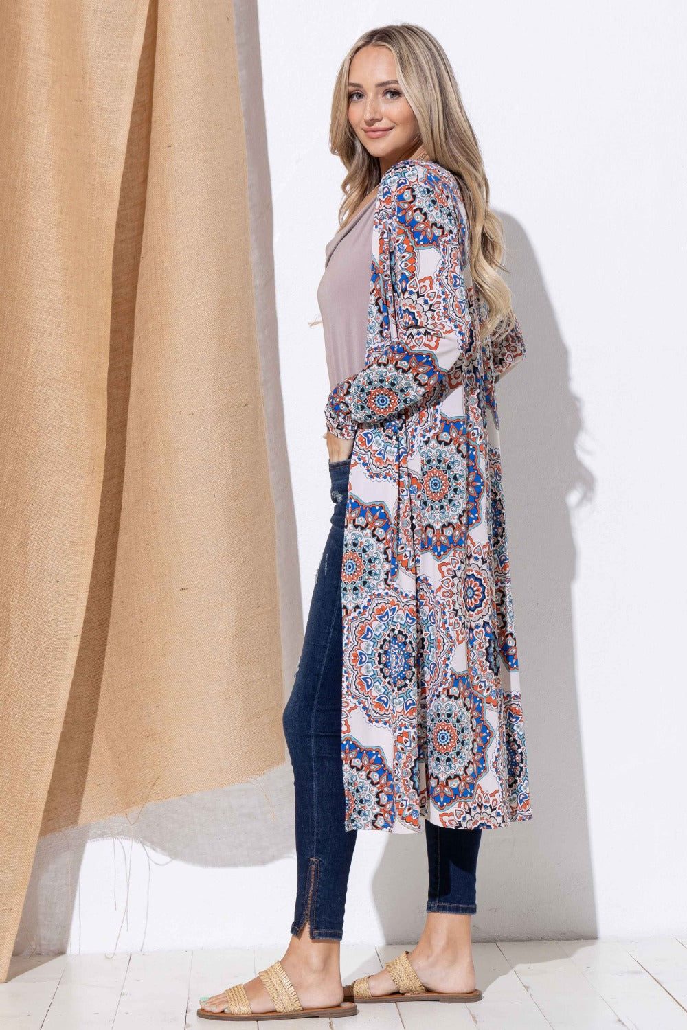 A woman wearing the "And The Why Printed Kimono Open Front Longline Cardigan" stands sideways, styled with a light top, dark jeans, and sandals. She smiles softly against a simple backdrop with a beige curtain.