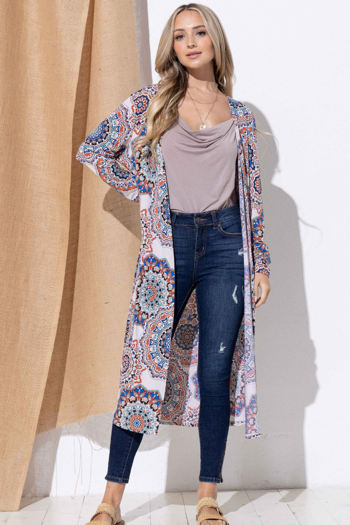 A person wearing the And The Why Printed Kimono Open Front Longline Cardigan over a light gray top and distressed blue jeans stands in front of a beige curtain. They have long hair, sandals, and a necklace, completing their effortlessly chic look.