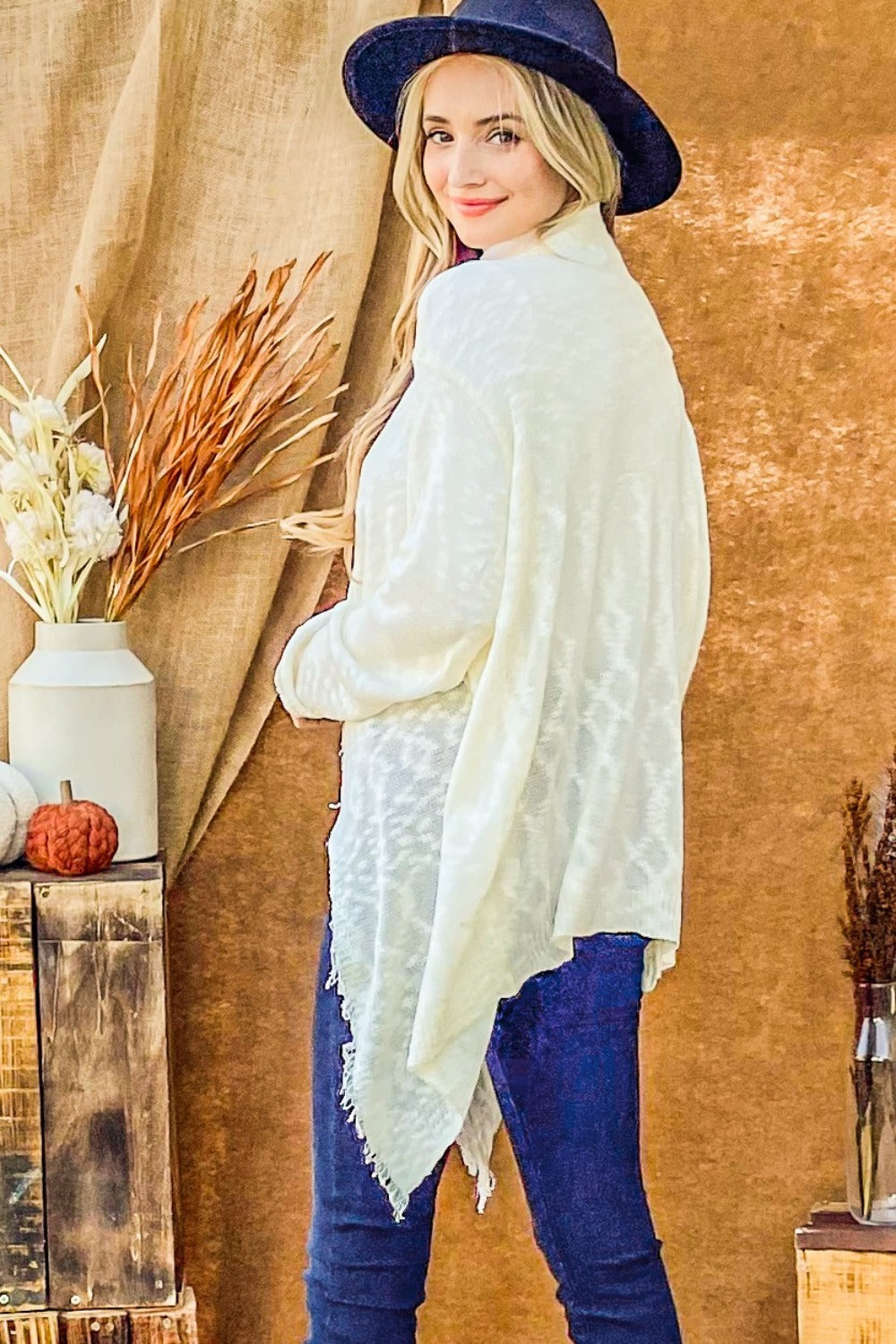 A person with long hair wearing a dark hat, an "And The Why Textured Knit Fringe Hem Cardigan," and jeans stands sideways in a rustic setting. The scene is adorned with autumn-themed decorations, including dried plants and a small pumpkin on wooden crates, adding to the bohemian flair.