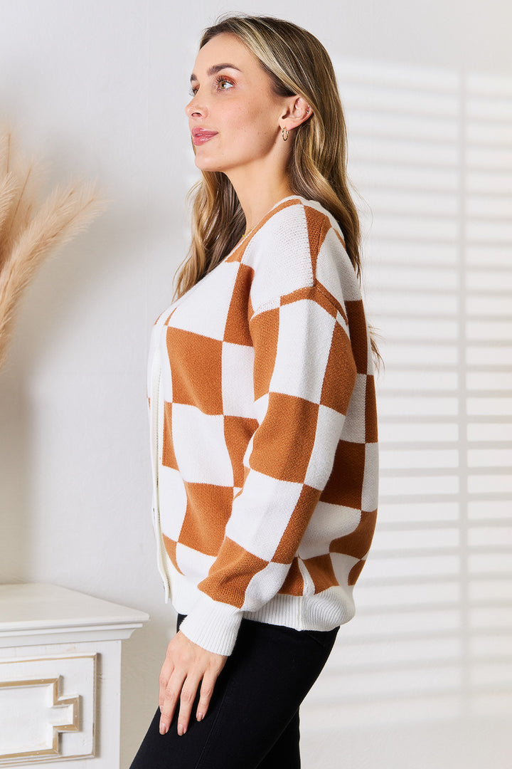 A person with long hair is standing sideways, wearing the cozy Angel Wings Double Take Button-Up V-Neck Dropped Shoulder Cardigan in a white and brown checkered pattern, paired with black pants. They're positioned next to a white wall that features window shadows and a small dresser decorated with pampas grass.