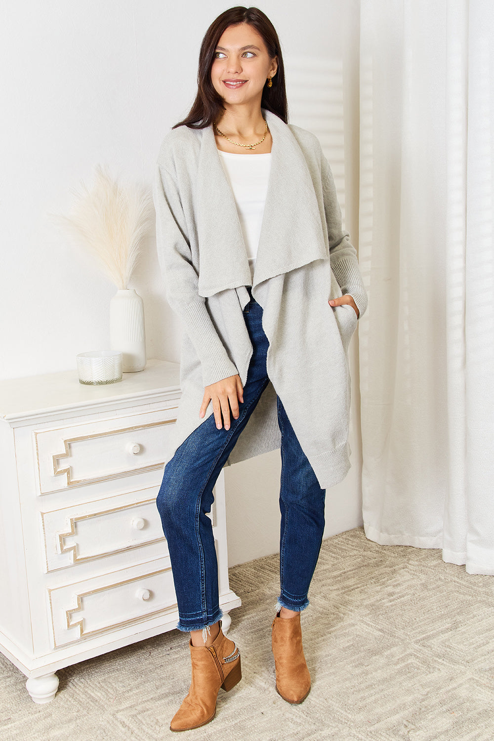 A woman stands smiling in a cozy room, embodying a cozy design. She wears a light gray Angel Wings Open Front Duster Cardigan with Pockets, complemented by a white top and blue jeans alongside tan ankle boots. Her right hand is tucked into her pocket as she leans slightly against a white dresser adorned with decorative items.