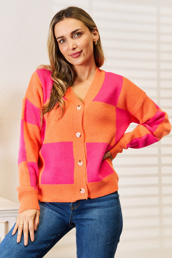 A person wearing the Angel Wings Woven Right Checkered V-Neck Dropped Shoulder Cardigan, featuring vibrant pink and orange patterns, stands indoors. With a smile on their face and long hair flowing down, they lean against a wall while dressed in jeans. This stylish cardigan adds a lively touch to their casual ensemble.