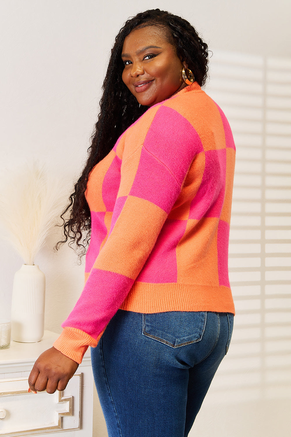 A person with long curly hair smiles while wearing the Angel Wings Woven Right Checkered V-Neck Dropped Shoulder Cardigan in vibrant pink and orange over blue jeans. They stand in a well-lit room beside a white table with a vase.