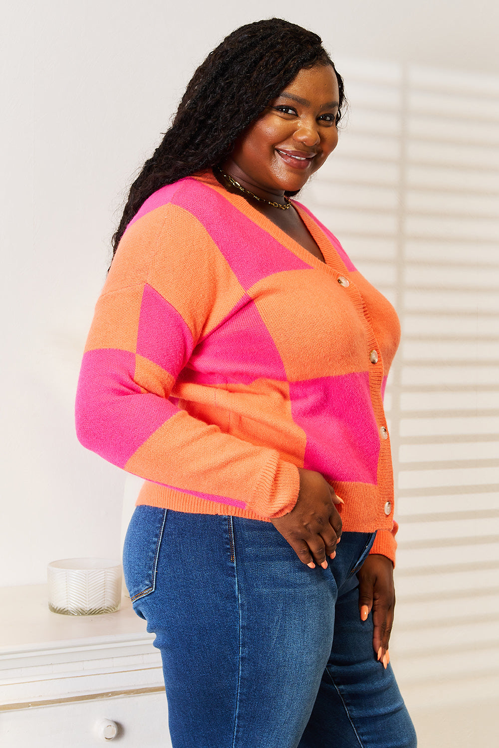 A woman smiles while wearing the Angel Wings Woven Right Checkered V-Neck Dropped Shoulder Cardigan in bright orange and pink, paired with blue jeans. She stands in a well-lit room featuring a light-colored wall and a small table in the background.