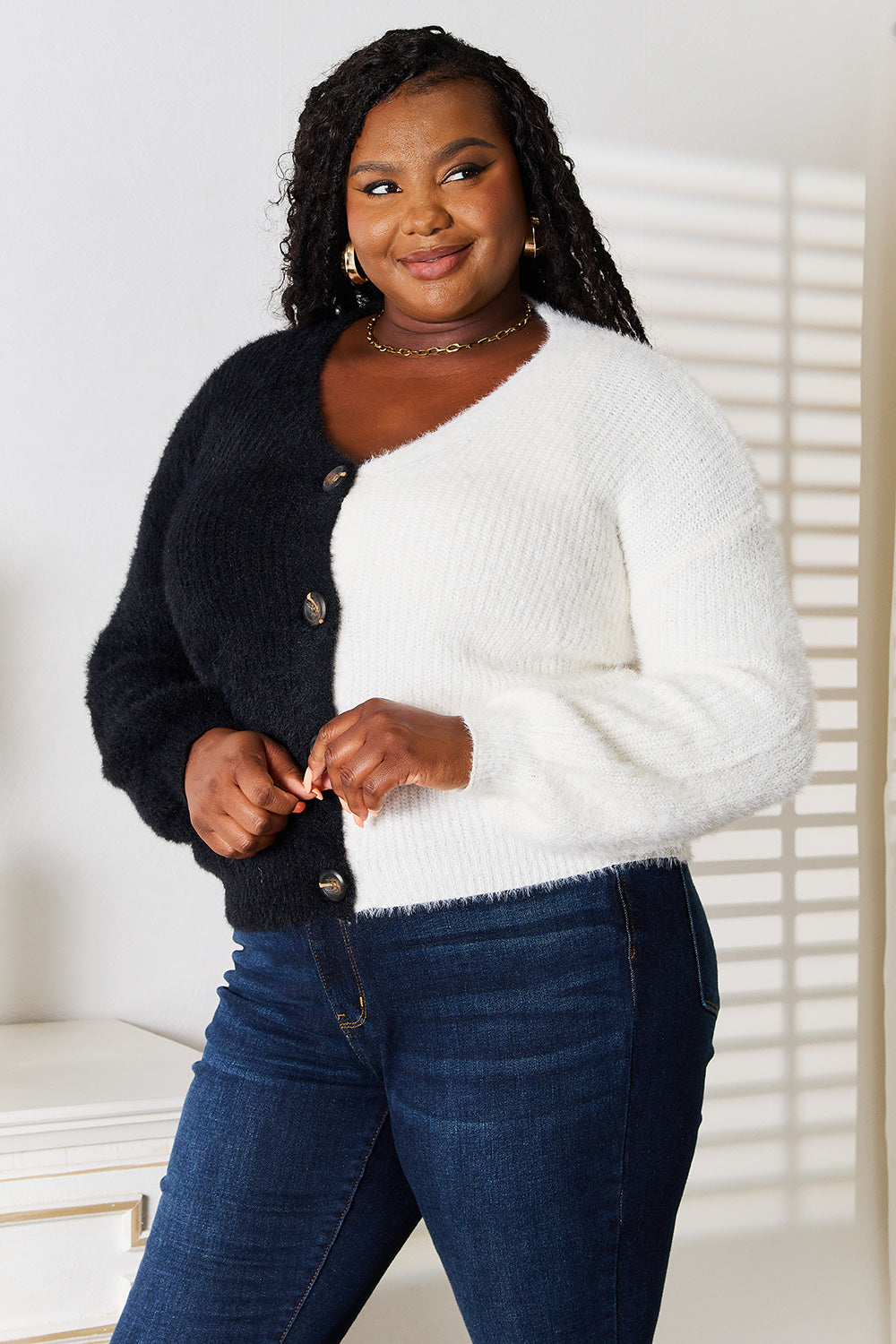 A person smiles while wearing the Angel Wings Woven Right Contrast Button-Front V-Neck Cardigan, a two-tone black and white garment made from stretchy material, paired with blue jeans. They stand in a well-lit room, where shadows from the blinds are cast on the wall.