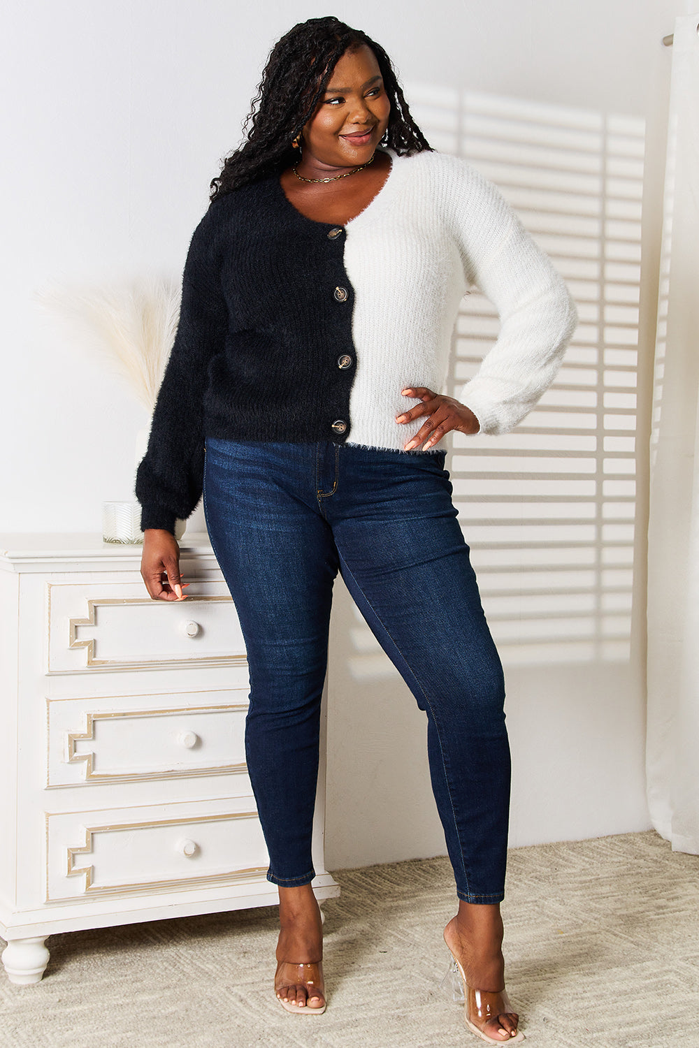 A person wearing the Angel Wings Woven Right Contrast Button-Front V-Neck Cardigan, featuring a half-black, half-white design, and blue jeans stands smiling in a room. They are in front of a white dresser adorned with decorative items. Sunlight casts striped shadows on the wall behind them, highlighting the contrast of their button-front attire.