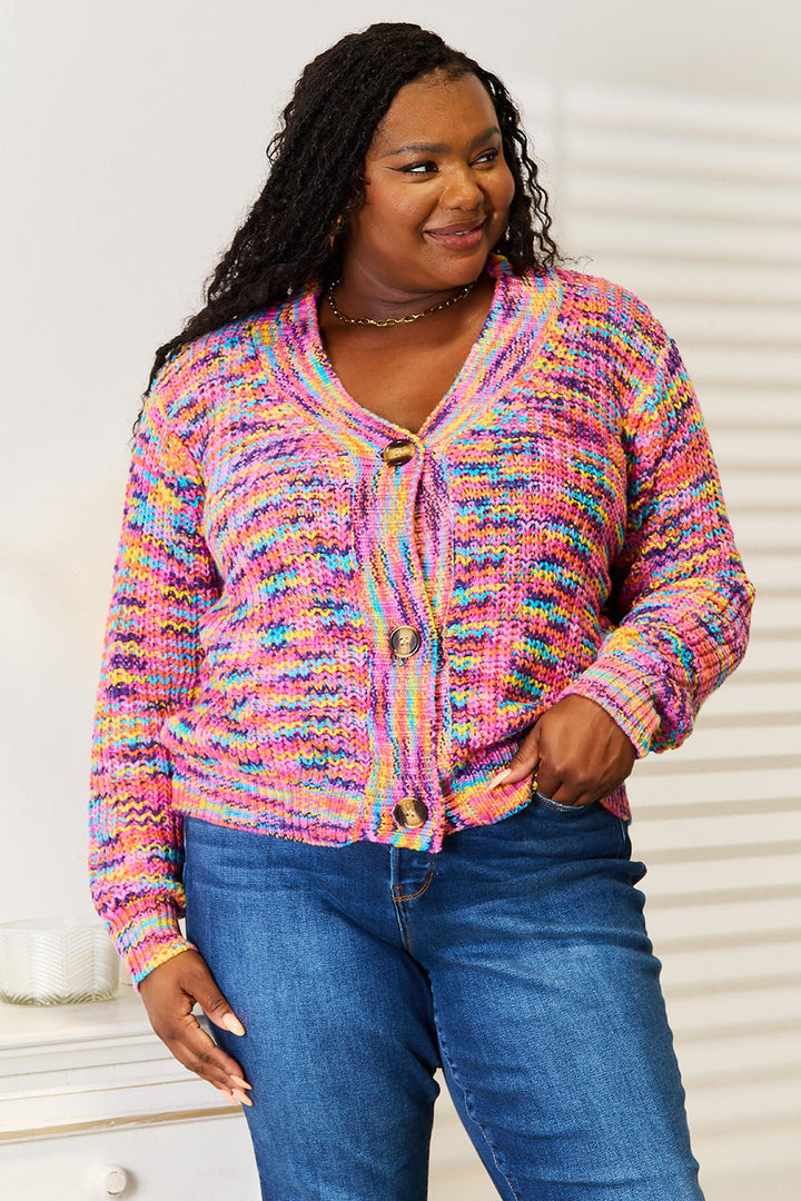 A person with long hair is wearing the Angel Wings Woven Right V-Neck Long Sleeve Cardigan, made of 100% acrylic featuring large buttons, paired with blue jeans. They are smiling and standing next to a white piece of furniture in a room with light walls.