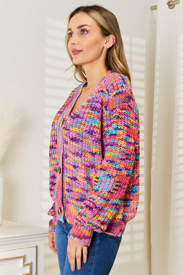 A woman wearing the Angel Wings Woven Right V-Neck Long Sleeve Cardigan stands in a softly lit room with white walls and subtle decor. Her long, straight hair cascades down as she gazes off to the side. The colorful, patterned cardigan made from 100% acrylic perfectly complements the serene ambiance.