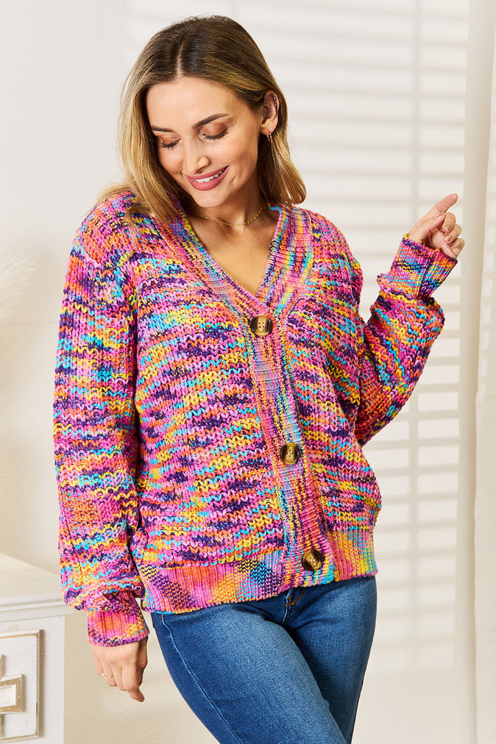 A person wearing the colorful Angel Wings Woven Right V-Neck Long Sleeve Cardigan, featuring chunky design elements and large wooden buttons, is standing in a well-lit room. With long hair and a smile, they are pointing to the right as window blind shadows create a pattern on the wall behind them.