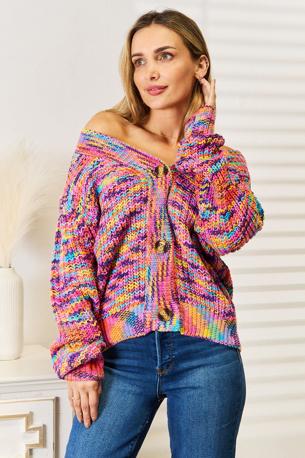 Indoors, a person wears the vibrant Angel Wings Woven Right V-Neck Long Sleeve Cardigan, featuring bold, bright patterns in an oversized knit. The cardigan's long sleeves and large buttons add to its charm as it's casually draped off one shoulder. Crafted from 100% acrylic, the individual softly touches their hair with one hand while pairing the look with blue jeans.