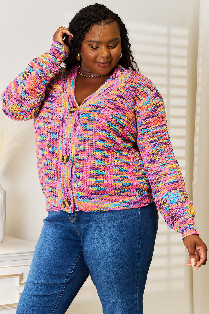 A person wearing the Angel Wings Woven Right V-Neck Long Sleeve Cardigan in multicolor and blue jeans stands in a bright room. They are looking down with a gentle smile, one hand touching their hair. Shadows from a window are visible on the wall, highlighting the 100% acrylic texture of the long-sleeve cardigan.