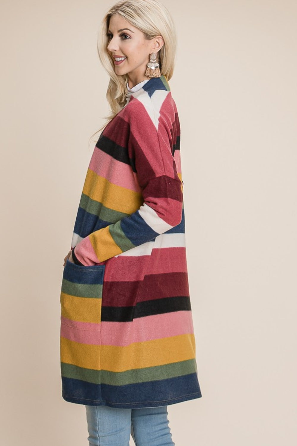 A woman with long blonde hair is wearing the BOMBOM Color Block Striped Open Front Cardigan, showcasing hues of white, red, pink, black, green, and mustard. She has paired it with blue jeans and large circular earrings while standing against a plain beige background.