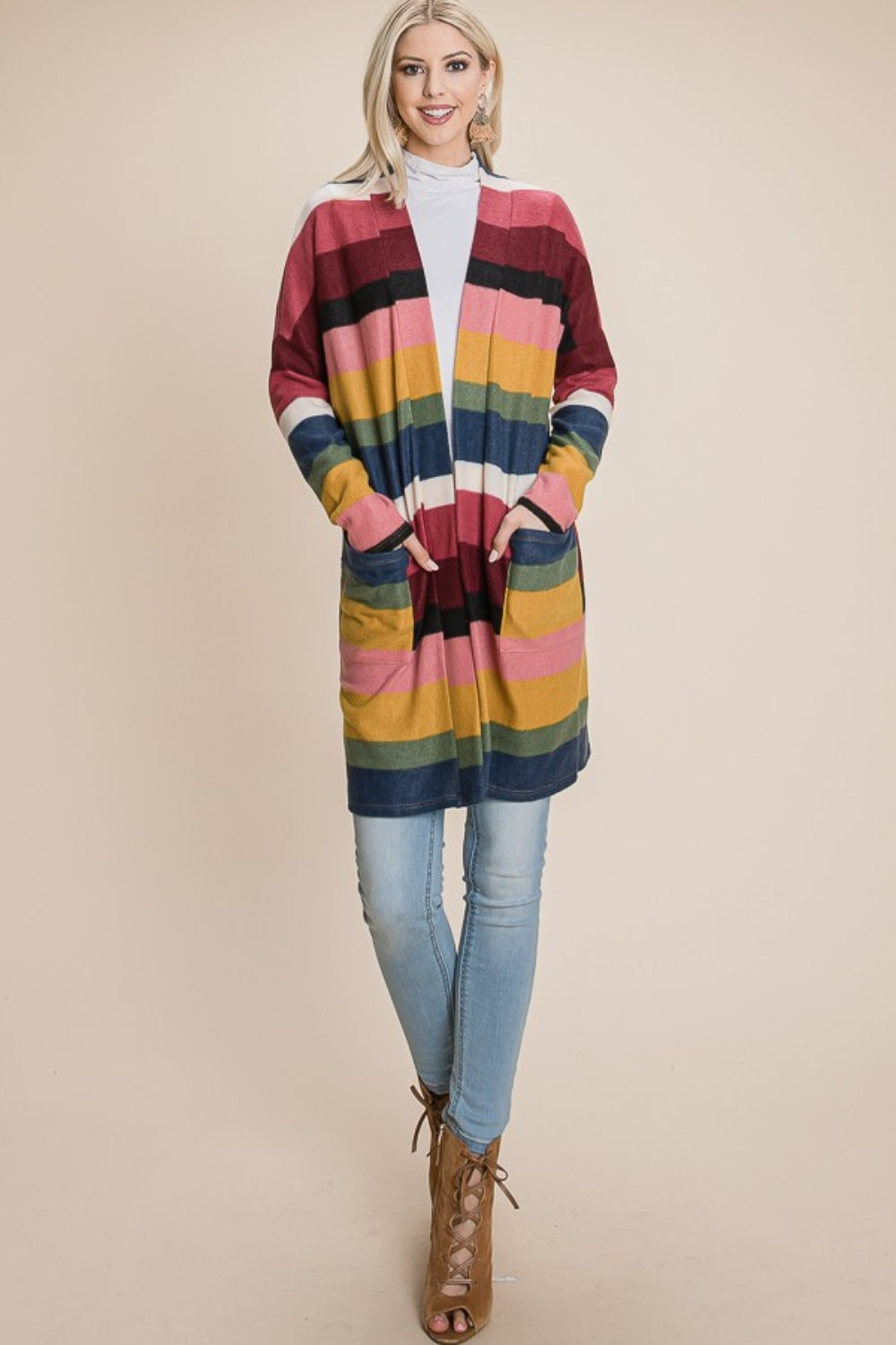 Against a beige background, a woman showcases the BOMBOM Color Block Striped Open Front Cardigan, featuring shades of pink, yellow, navy, and green. She pairs it with light blue jeans and tan ankle boots and smiles with her hands in the cardigan pockets.