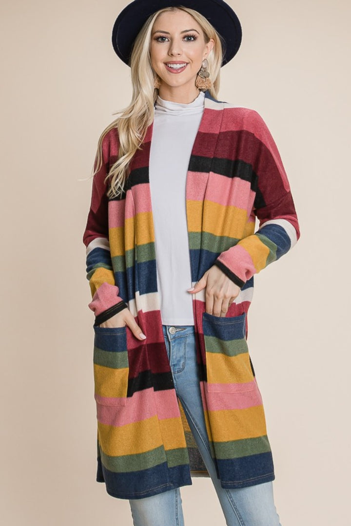 A person in a wide-brimmed hat wears the BOMBOM Color Block Striped Open Front Cardigan, showcasing its long and versatile design over a white top and blue jeans. This cardigan features vibrant stripes in shades of pink, yellow, blue, and red against a plain beige background.