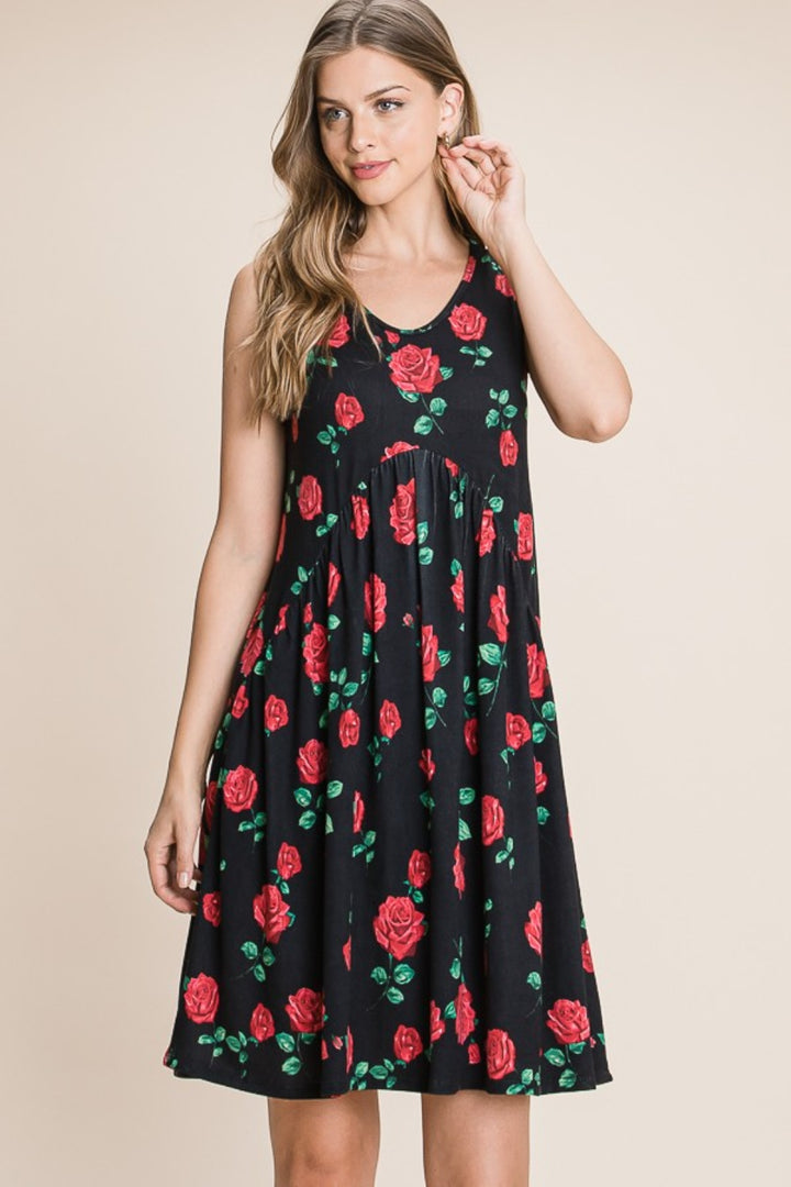 Wearing the BOMBOM Floral Ruched Tank Dress in sleeveless black with red rose patterns, a person stands against a neutral background. They have long wavy hair and touch their ear with one hand while the other arm rests by their side.