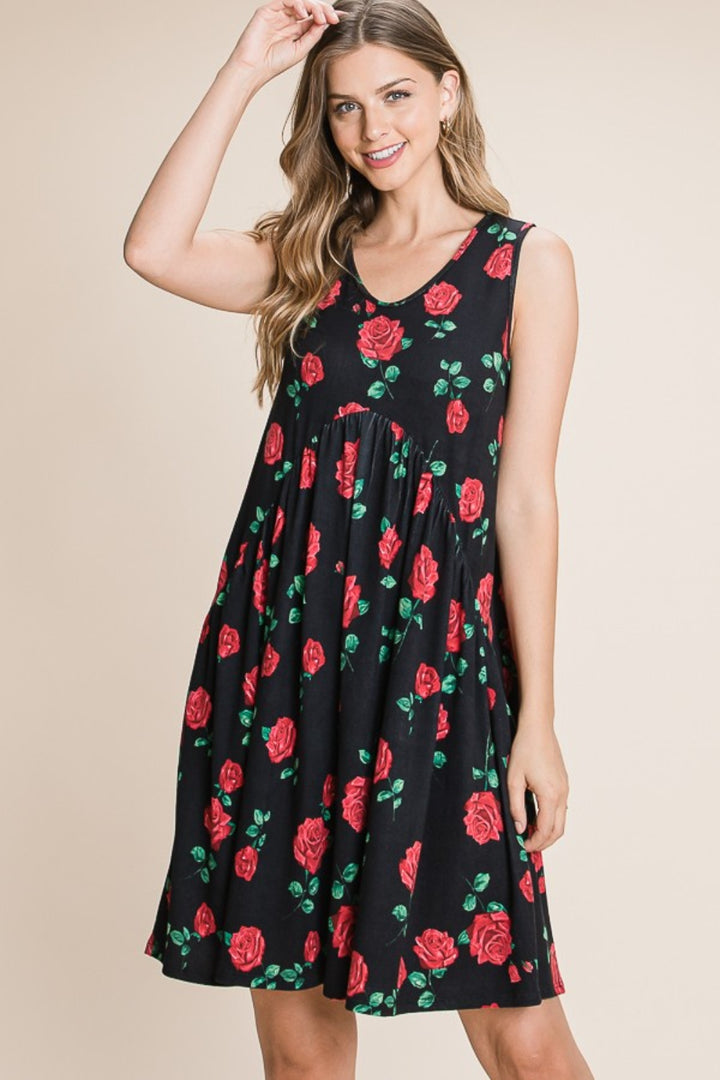 A woman wears a sleeveless BOMBOM Floral Ruched Tank Dress with red roses and green leaves. Her long, wavy hair flows as she smiles, gently touching her head against a simple beige backdrop that highlights the beauty and versatility of her ensemble.