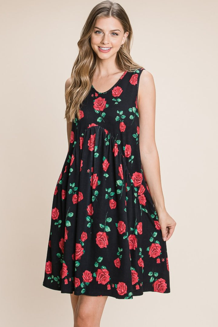 A smiling woman with long hair models the BOMBOM Floral Ruched Tank Dress, a sleeveless black fabric with vibrant red roses and green leaf patterns, against a simple beige backdrop.