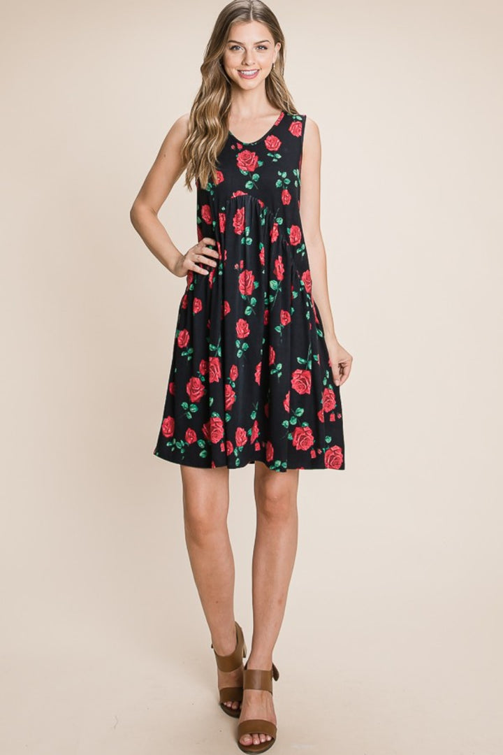 A person smiles in front of a beige background, wearing a BOMBOM Floral Ruched Tank Dress with red rose patterns, brown sandals, and has long wavy hair.