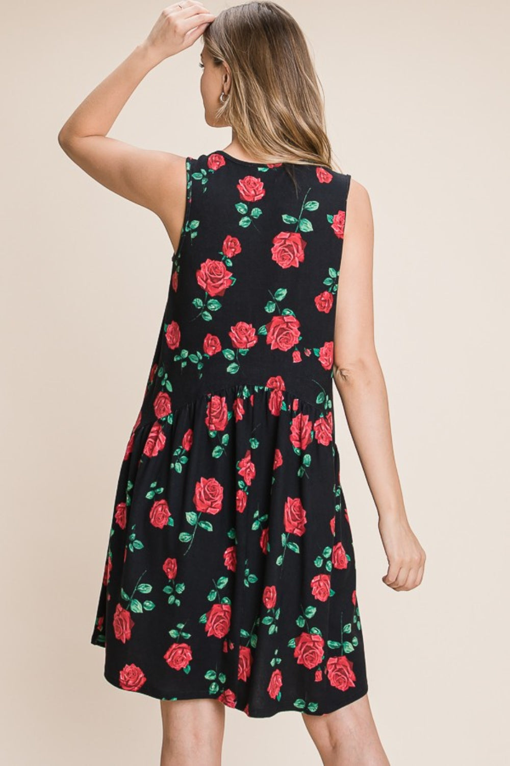 A woman poses against a plain beige background, adjusting her hair while wearing the BOMBOM Floral Ruched Tank Dress, a sleeveless black piece with vibrant red roses and green leaves.