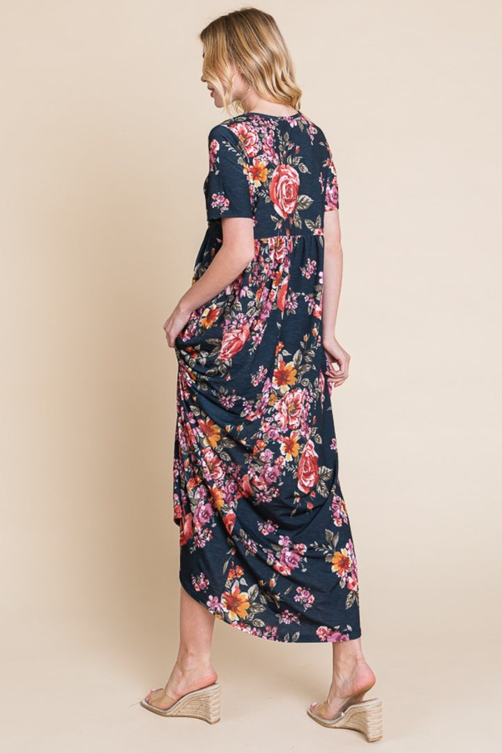 A person with shoulder-length hair wears a navy BOMBOM Floral Short Sleeve Maxi Dress decorated with pink, red, and orange flowers. Against a beige background, they look to the side in wedge sandals, highlighting the dresss versatile flowy silhouette.