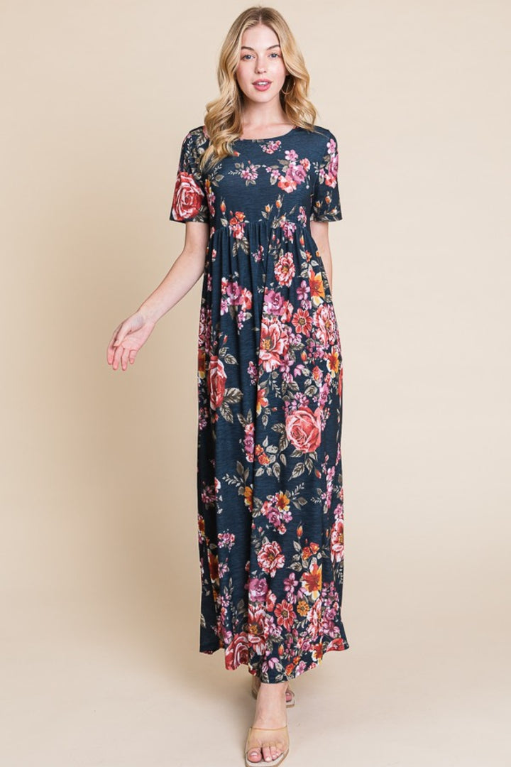 A woman stands against a beige background wearing the BOMBOM Floral Short Sleeve Maxi Dress with a flowy silhouette, adorned with pink, red, and orange flowers. Her loose blonde waves complement the versatile look, completed with neutral sandals.