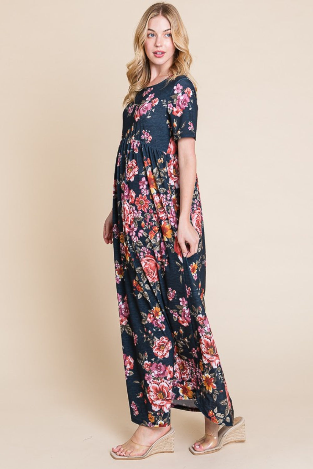 A woman in the BOMBOM Floral Short Sleeve Maxi Dress, boasting a versatile flowy silhouette and dark floral design, poses against a beige background. Turning sideways, she gently smiles at the camera while her wedge sandals elegantly elevate her style.