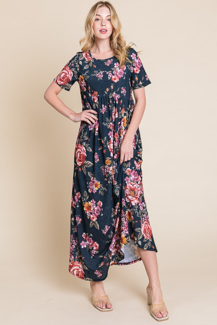 Against a beige background, a woman poses confidently in the BOMBOM Floral Short Sleeve Maxi Dress, featuring black fabric adorned with pink and red floral accents. Her long blonde hair falls gracefully as she stands in open-toed beige heels, with one hand on her hip.