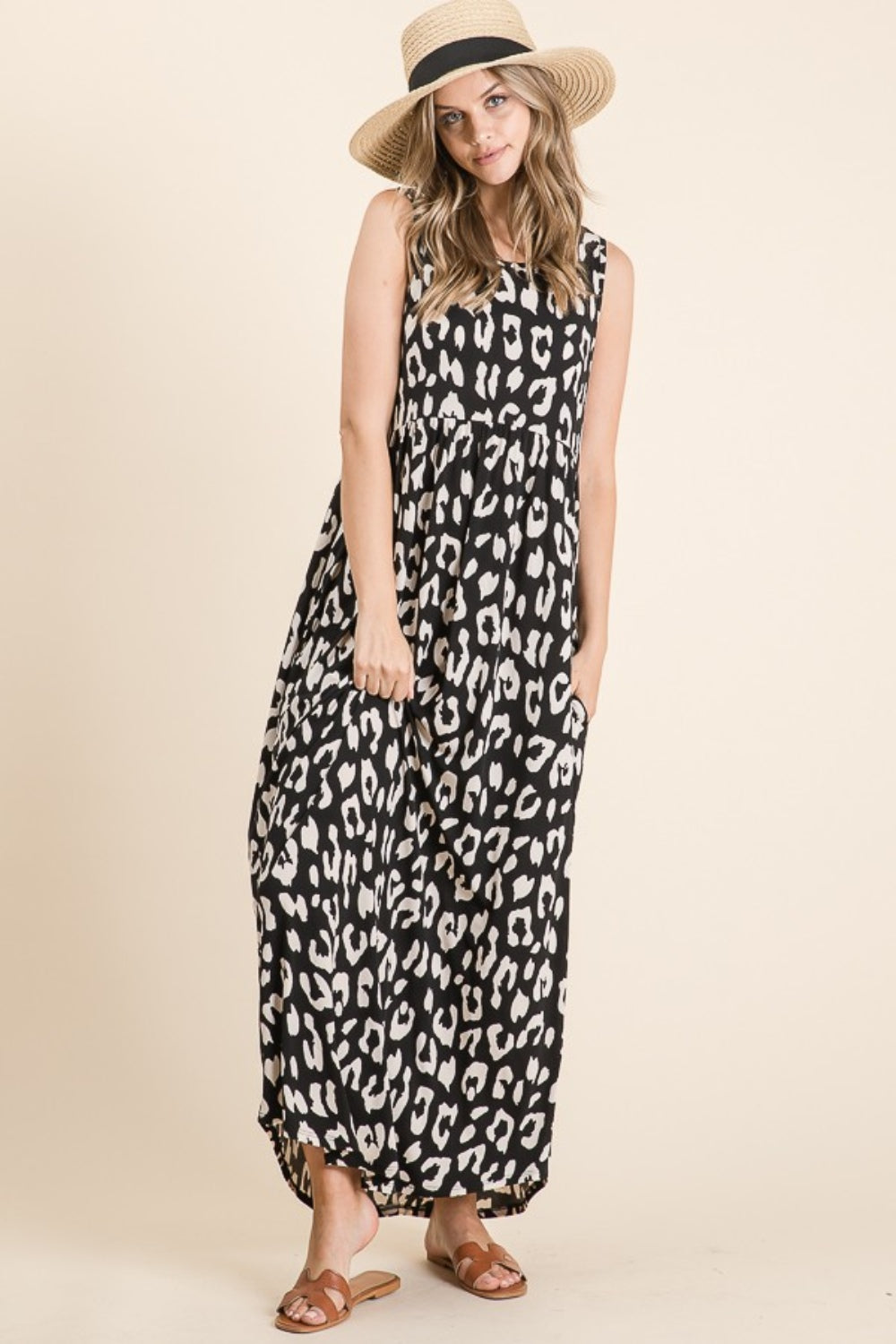 A woman in a BOMBOM Leopard Maxi Dress with pockets and a straw hat stands against a plain background. Her long hair flows freely, and she wears brown sandals, her hands casually resting in the shirring waist of the black and white dress.